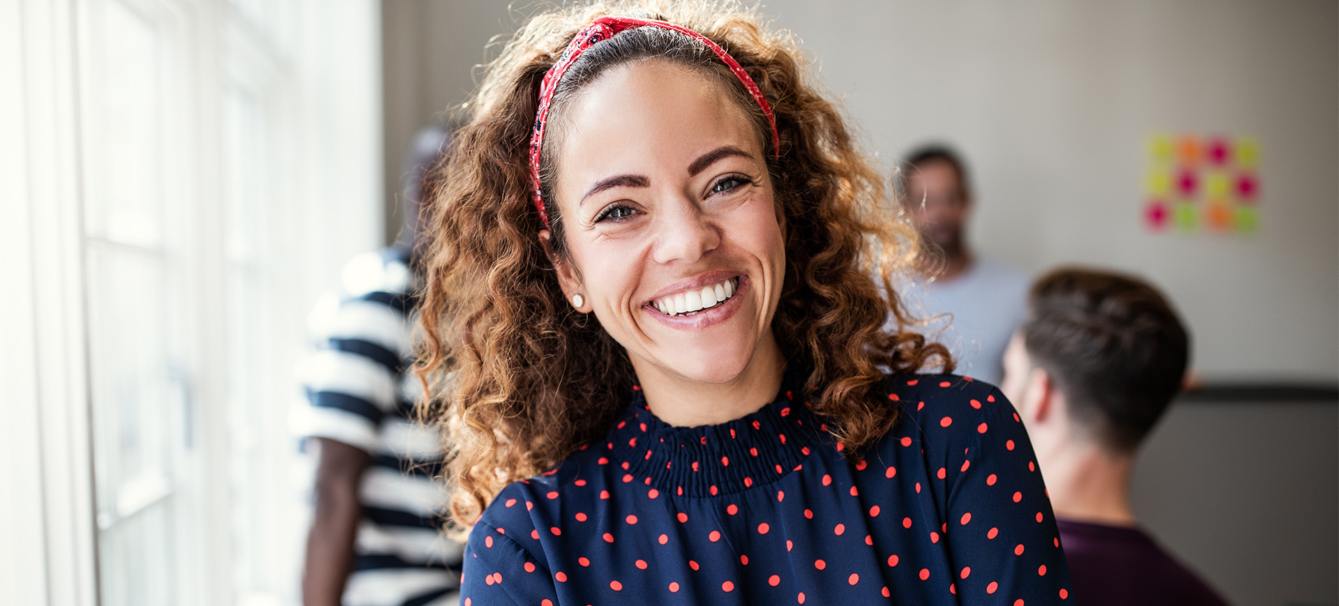 Woman with curly hair in blue shirt with red dots smiling next to sunny window